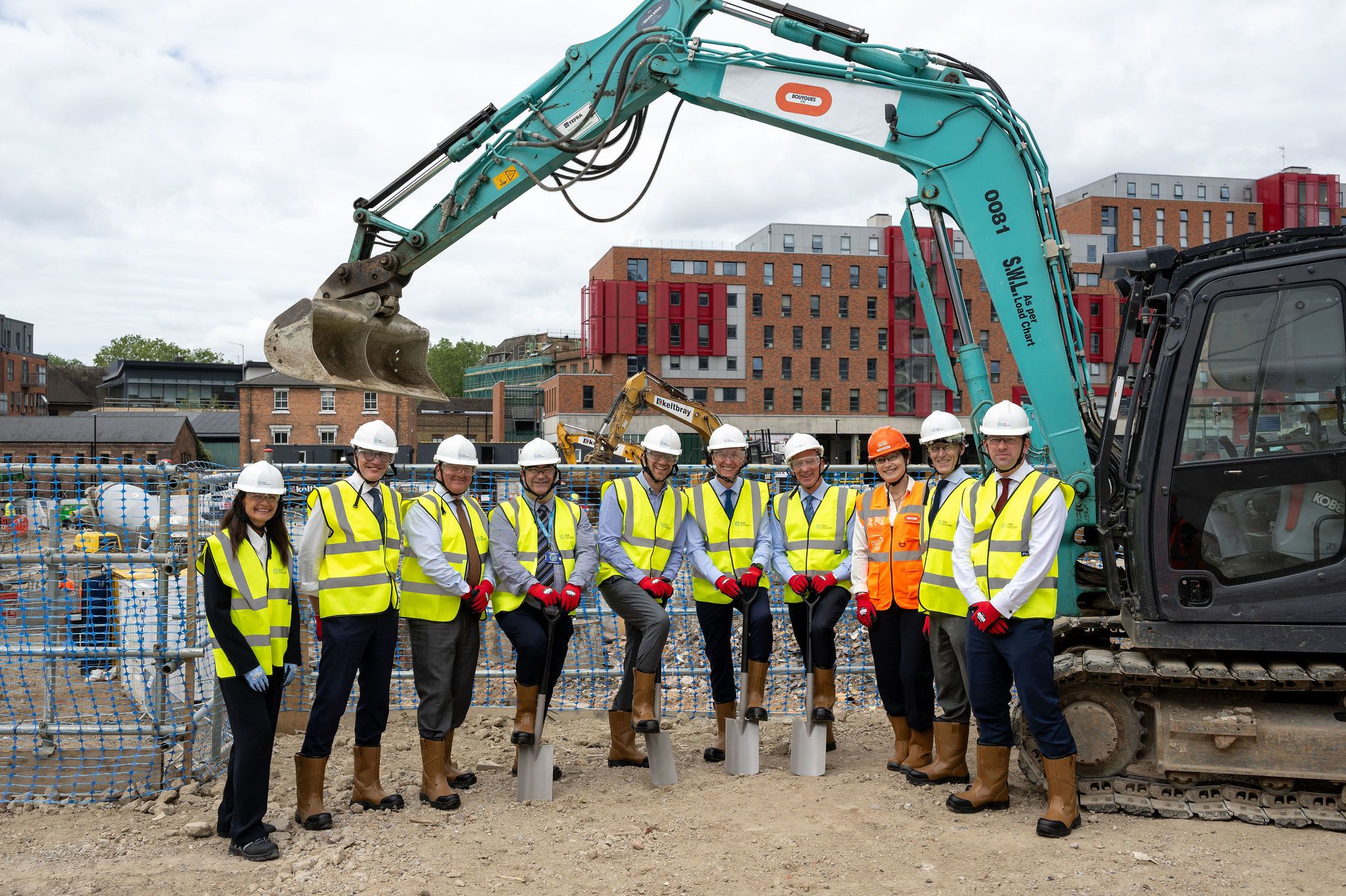 A group in hard hats and high vis vests, some holding spades, stand in front of a digger on a building site