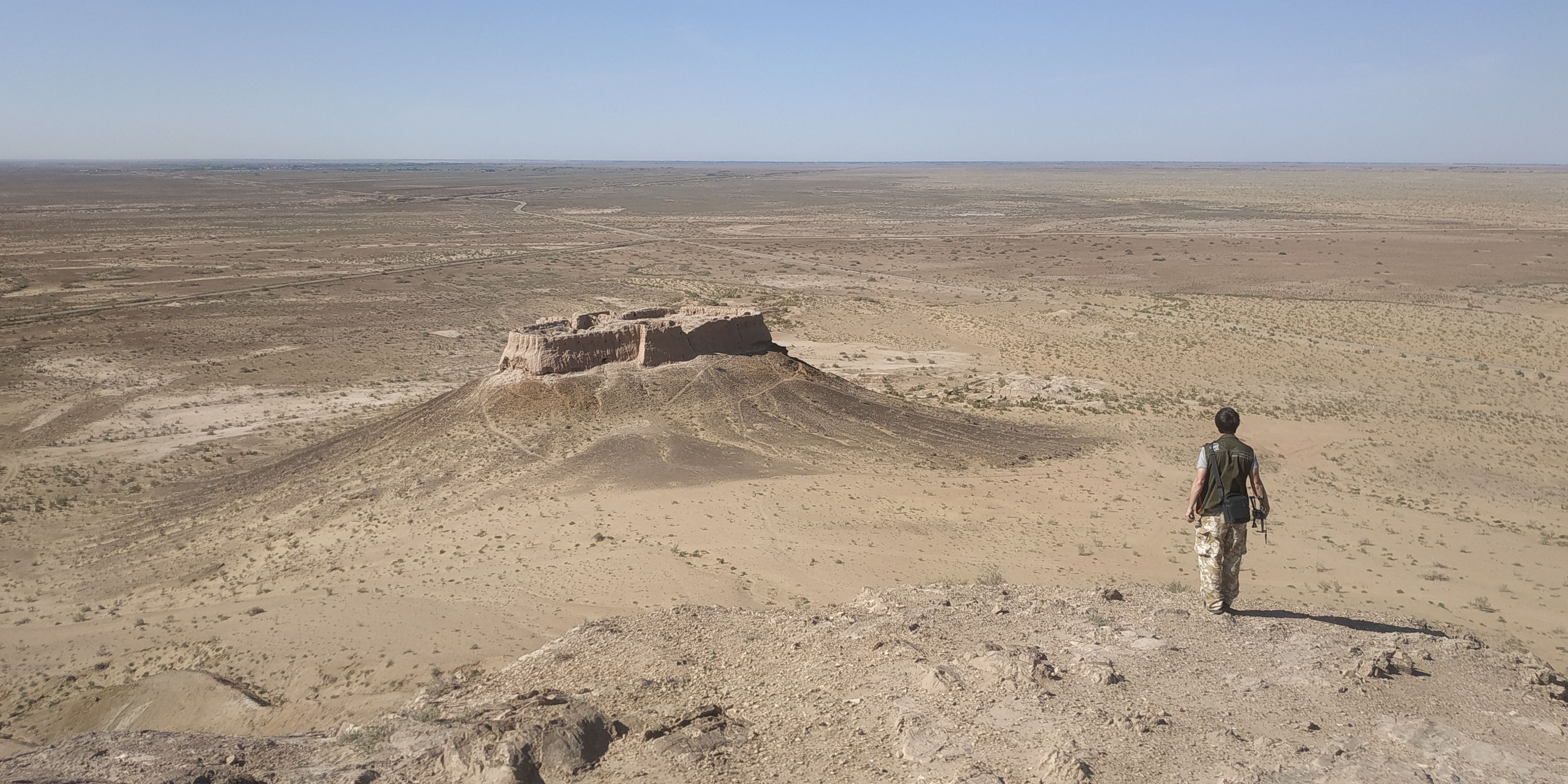 Foreground: a researcher walks downhill in a sandy environment. Background: a large historic cultural site on a mound, against an empty backdrop.